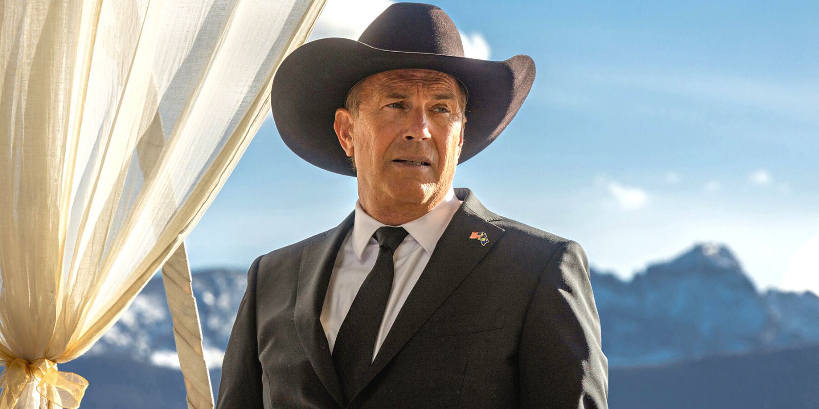 Kevin Costner con un traje negro y un sombrero de diez galones, parado frente a una cadena montañosa